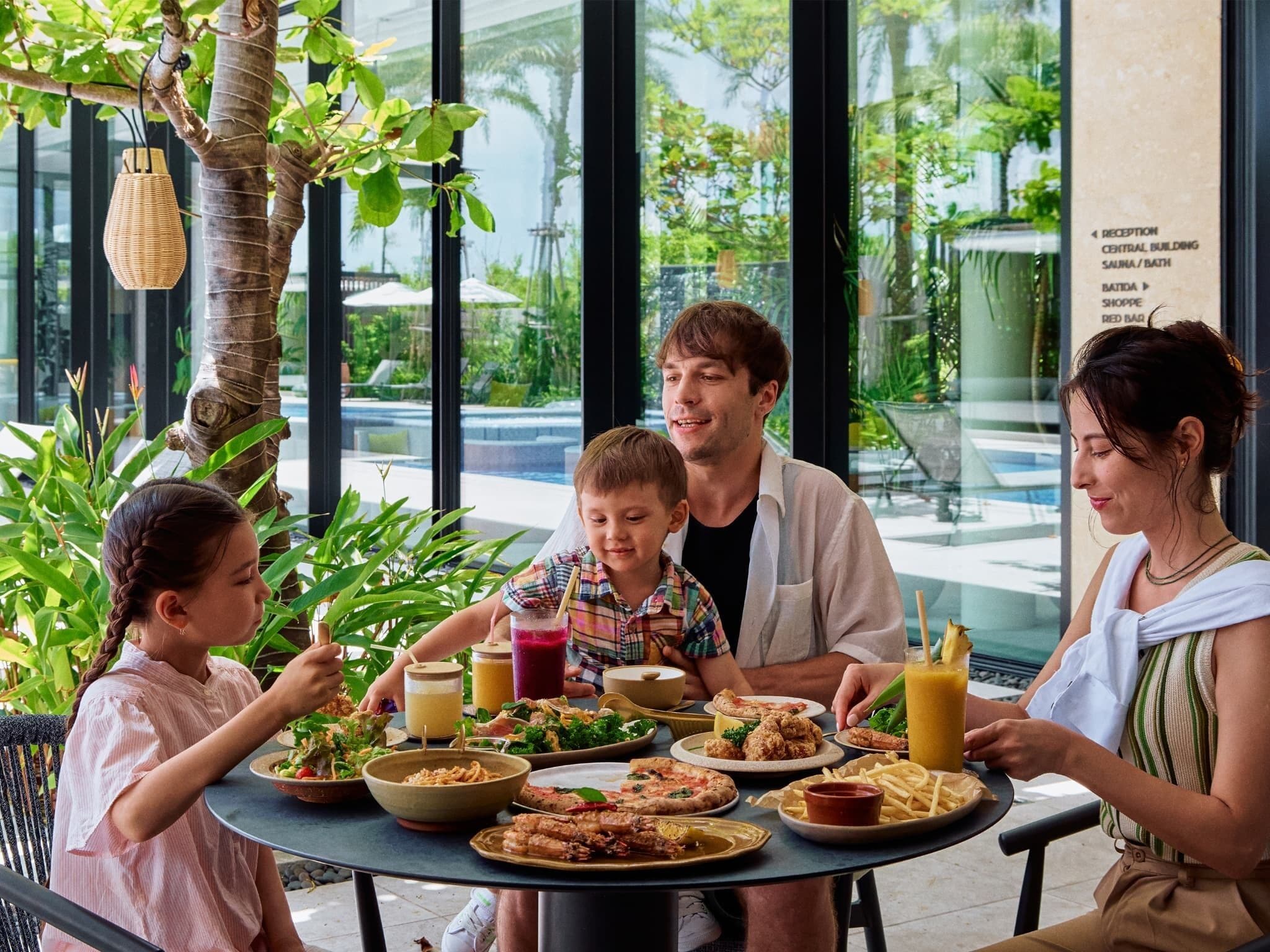 A family dining at an outdoor table with various dishes and drinks, surrounded by greenery and large windows.