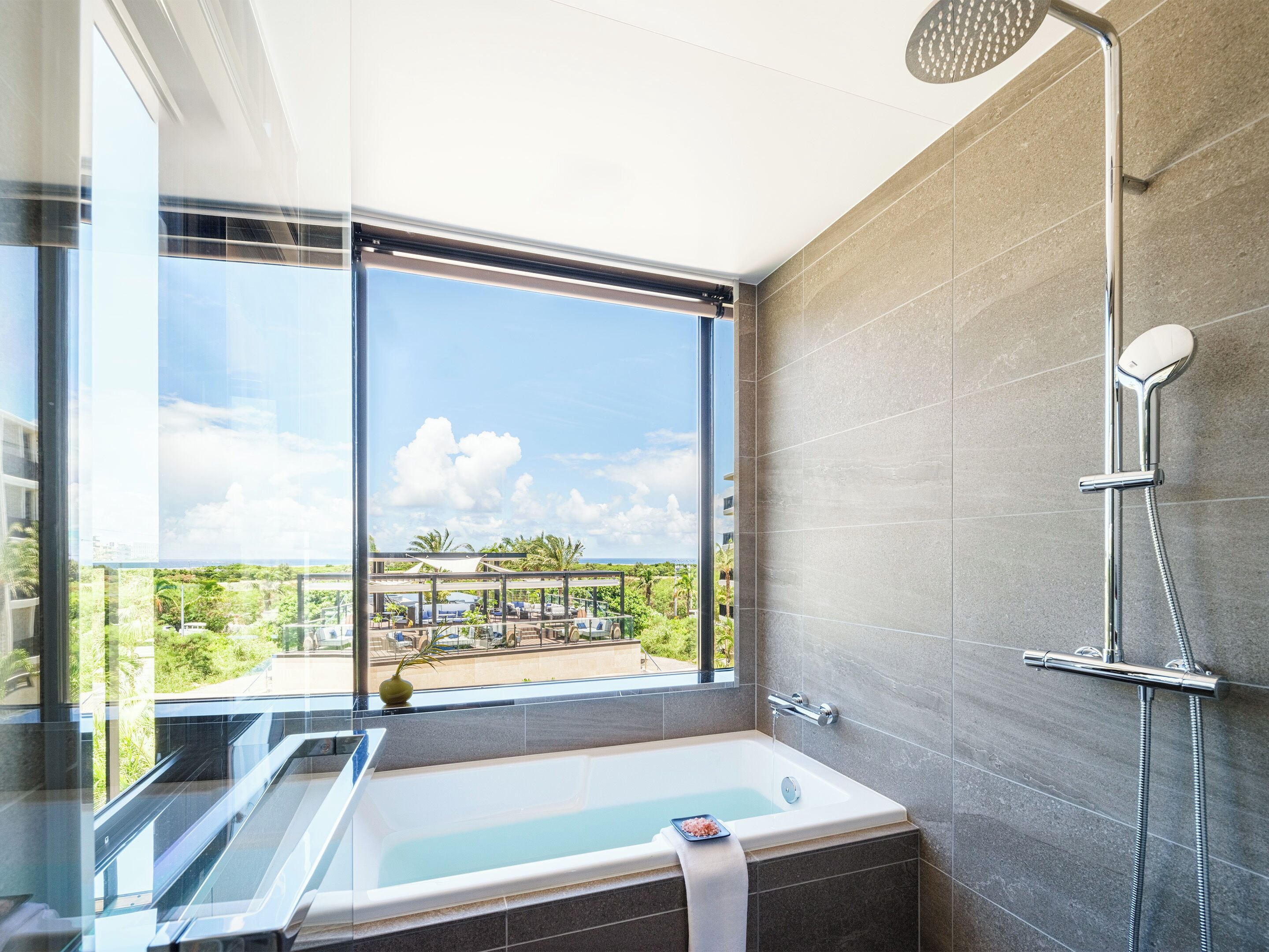 A modern bathroom with a bathtub, shower, and a large window offering a view of a pool and greenery.