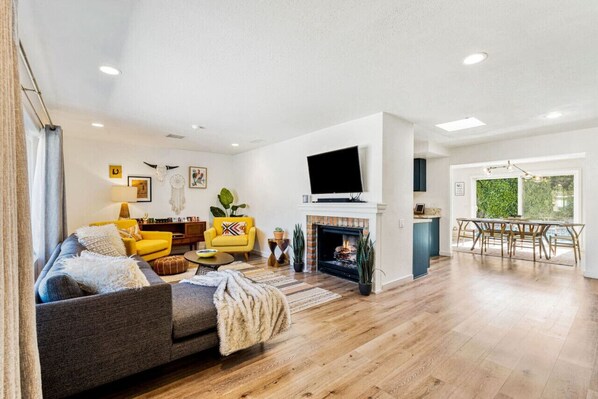 A spacious living room with a fireplace, a yellow armchair, a grey sofa, and a dining area with a table and chairs. - Modern Desert Oasis with Sweeping Mountain Views (Palm Springs)