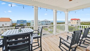 Eine überdachte Terrasse im Freien mit Holzboden und weißen Geländern, auf der ein Tisch und Stühle stehen, mit Blick auf eine Küstenlandschaft mit Häusern und dem Meer.