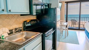 Cozinha com fogão, pia e armários, área de jantar com mesa e cadeiras, e varanda com vista para o oceano.