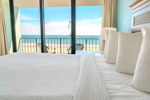 A hotel room with a bed, a view of the beach, and a balcony with chairs.