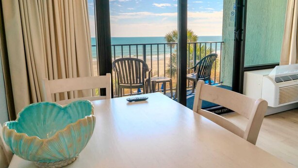Um quarto com uma mesa, cadeiras, uma tigela decorativa e vista para a praia.
