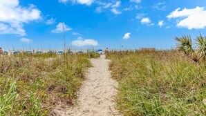 Um caminho de areia margeado por grama, que leva ao mar sob um céu claro.