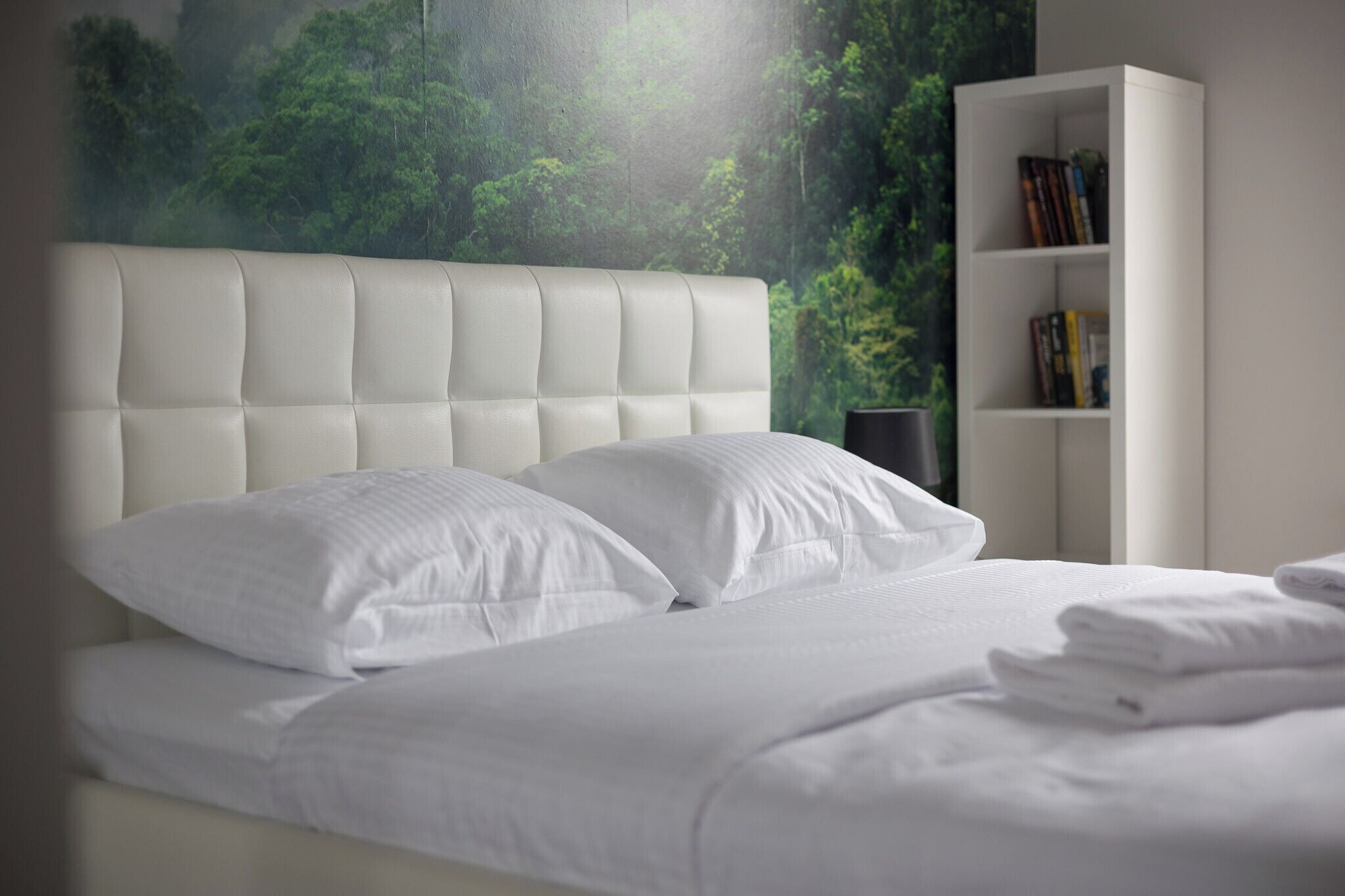 A neatly made bed with white linens and pillows, a white headboard, and a bookshelf with books.