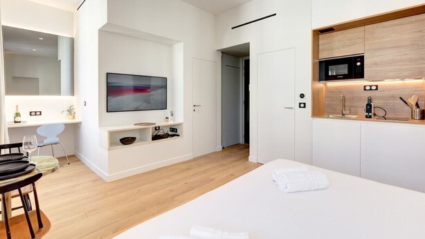 A modern hotel room with a large bed, a flat-screen TV mounted on the wall, a wooden floor, and a kitchenette with a microwave. - Upstays Renovated 27m² Studio Near Palais (Cannes)