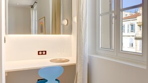 A room with a white desk, a blue chair, a window with a view of buildings, and a mirror reflecting the room. - Upstays Renovated 27m² Studio Near Palais (Cannes)
