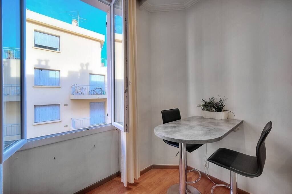 Una habitación pequeña y moderna con una mesa redonda y dos sillas, una planta en maceta y vistas a edificios vecinos a través de una ventana.