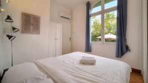 A bedroom with a bed, a bedside lamp, a window with curtains, and an air conditioning unit. - Upstays Garden Terrace 74m² Apartment (Cannes)