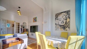 A modern living room with a sofa, a dining table with yellow chairs, a TV, and a large abstract painting on the wall. - Upstays Garden Terrace 74m² Apartment (Cannes)