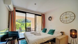 A hotel room with a bed, a desk, a chair, a standing lamp, and a view of greenery outside.