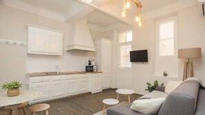 Een moderne keuken met witte kastjes, een eethoek met krukken, een grijze bank en een aan de muur bevestigde televisie.