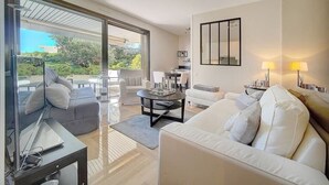 A modern living room with a sofa, armchairs, a coffee table, and a dining area with a table and chairs. There is a sliding glass door leading to an outdoor area with greenery. - Upstays Sea View Terrace 47m² Apartment (Cannes)