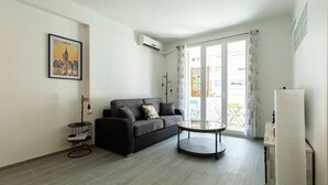 Een moderne woonkamer met een bank, een salontafel en een bijzettafeltje. Er hangt een foto aan de muur, een airconditioning en een raam met gordijnen.