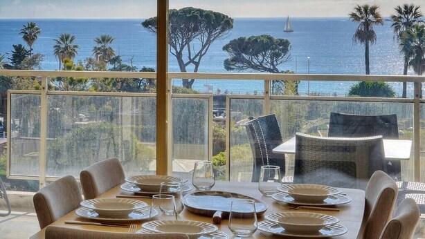 A dining area with a table set for six, overlooking a scenic ocean view with palm trees and a sailboat. - Upstays Sea View Pool Terrace 75m² Apartment (Cannes)