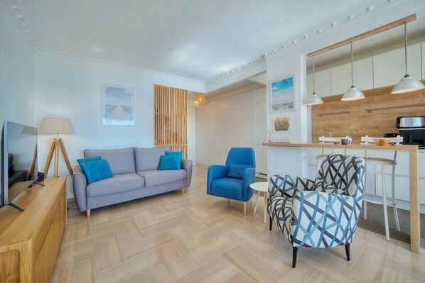 Un salon moderne avec un canapé gris, des fauteuils bleus et un fauteuil à motifs. On peut également apercevoir un meuble TV en bois et une petite table avec une lampe.