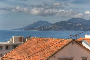 A coastal town with terracotta-roofed buildings, a view of the sea, and mountains in the background.