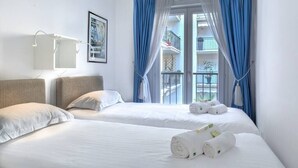 Une chambre d’hôtel avec deux lits, une grande fenêtre avec des rideaux bleus, un mur blanc et un tableau encadré.