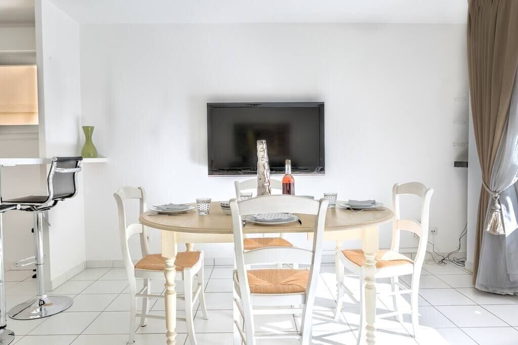 Een eetgedeelte met een ronde tafel, witte stoelen, een flatscreen televisie aan de muur en een kleine groene vaas op een plank.