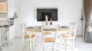 Un coin repas avec une table ronde, des chaises blanches, un téléviseur à écran plat fixé au mur et un petit vase vert posé sur une étagère.