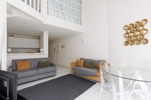 A modern living space with a grey sofa, a smaller sofa, a glass dining table, and a kitchen area.