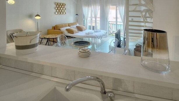Una habitación de hotel moderna con una cama grande, una zona de estar con sofá y una mesita, un baño con lavabo y un espejo amplio, y un balcón con barandilla.