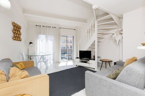 Ein modernes Wohnzimmer mit einem grauen Sofa, einem Holztisch, einem Fernseher auf einem weißen Schrank und einer Treppe, die zu einer oberen Etage führt.