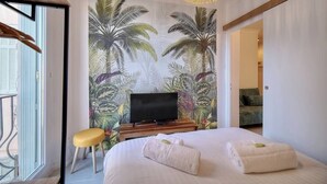 A bedroom with a large bed, a TV on a wooden stand, a yellow stool, and a tropical wallpaper. - Upstays cosy 20m² Apartment Suquet (Cannes)