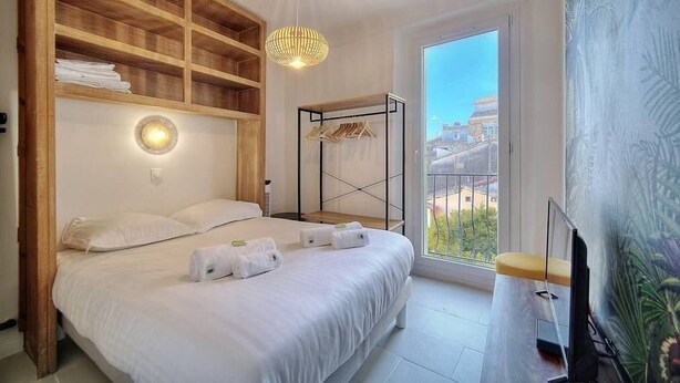 A bedroom with a bed, wooden shelves, a hanging light, a balcony with a view, and a painting. - Upstays cosy 20m² Apartment Suquet (Cannes)