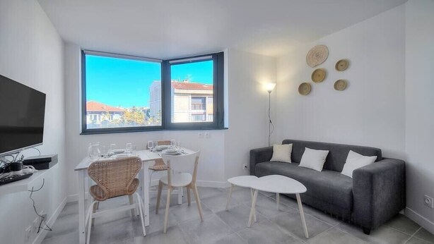 A modern living room with a dining area, a grey sofa, and a large window offering a city view. - Immogroom - Charming apartment - Close to beaches (Cannes)