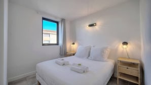 A neatly arranged bedroom with a large bed, two bedside tables, and a window with a view of buildings. - Immogroom - Charming apartment - Close to beaches (Cannes)