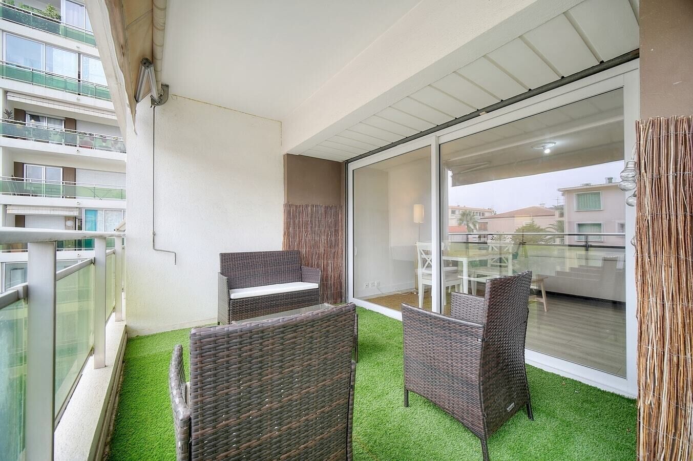Een balkon met rieten meubels en kunstgras, met uitzicht op een woonwijk.