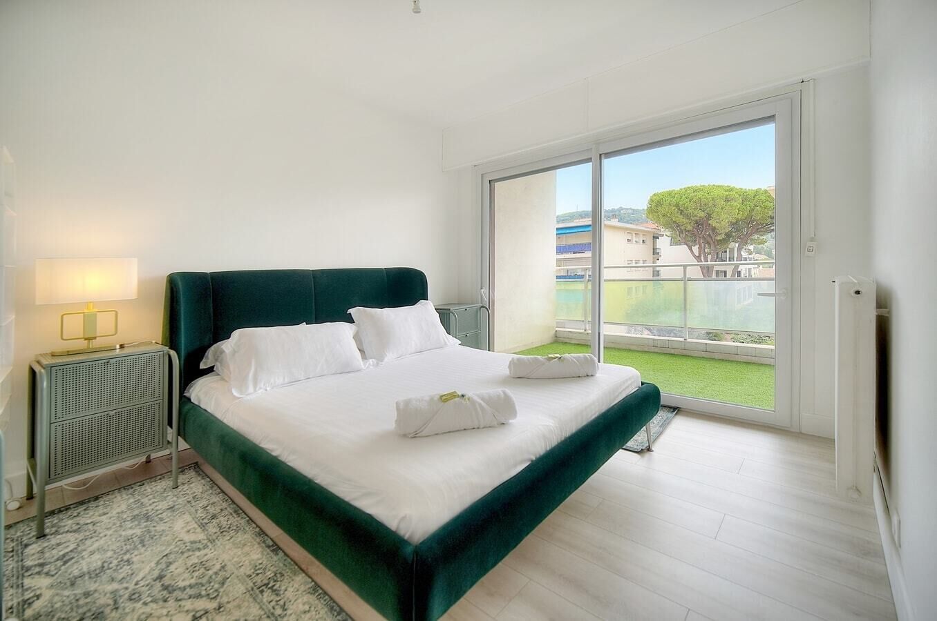 Ein Schlafzimmer mit einem großen Bett, einem Nachttisch, einer Lampe und Blick auf eine grüne Wiese sowie Gebäude durch eine Glastür.