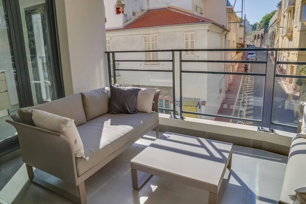 A balcony with a sofa and a table, overlooking a street with buildings and a red-roofed house.