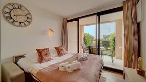 A bedroom with a large bed, a wall clock, a sliding glass door to a balcony, and a small table with a cactus. - Upstays Terrace Pool 37m² Apartment (Cannes)