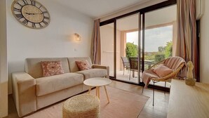 A living room with a sofa, a round table, a wicker ottoman, a wicker chair, a large window, and a wall clock. - Upstays Terrace Pool 37m² Apartment (Cannes)