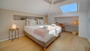 A bedroom with a large bed, a skylight, a bedside table with a lamp, and a small side table.