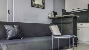 Een moderne woonkamer met een grijze bank, een klein rond tafeltje en een keuken in de achtergrond.