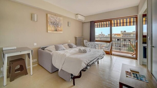 A modern hotel room with a bed, a small desk, a chair, and a balcony with a city view. - Upstays Terrace 25m² Studio Carnot (Cannes)