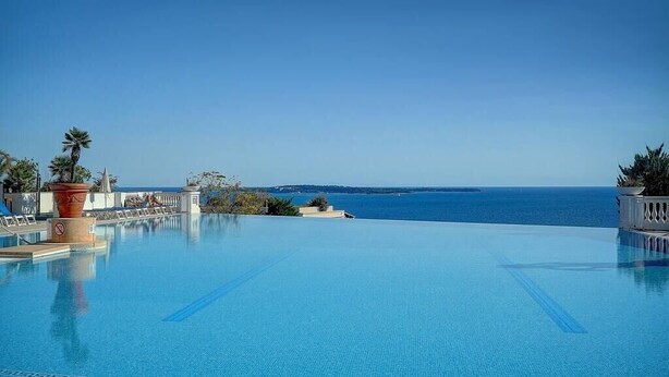 A large infinity pool with clear blue water, surrounded by a white fence, palm trees, and a view of the ocean. - Upstays Garden Terrace Pool 30m² Apartment (Cannes)