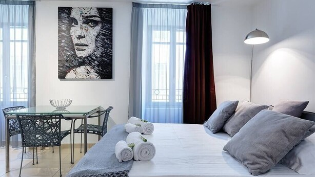 A hotel room with a bed, a desk, and a chair. There is a painting on the wall and a window with curtains. - Upstays Port Near 45m2 Apartment AC (Cannes)