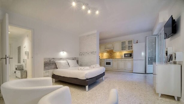 Une chambre moderne avec un lit, deux fauteuils blancs, un coin cuisine et un téléviseur fixé au mur.