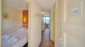 A hotel corridor with a bed on the left, a door with the number 1198, and a view of the sea through an open door at the end.