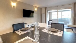 A modern hotel room with a dining table set for two, a flat-screen TV mounted on the wall, and a large window offering a view of the city. - Upstays Croisette Veranda 48m² Apartment (Cannes)