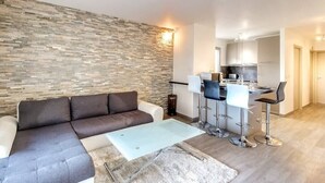 A modern living room with a sectional sofa, a glass coffee table, and a stone accent wall. A kitchen area with white cabinets and a dining space with bar stools is visible in the background. - Upstays Croisette Veranda 48m² Apartment (Cannes)