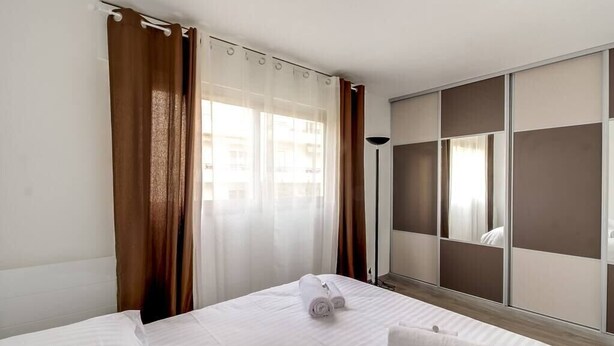 A hotel room with a bed, curtains, a wardrobe, and a floor lamp. - Upstays Croisette Veranda 48m² Apartment (Cannes)