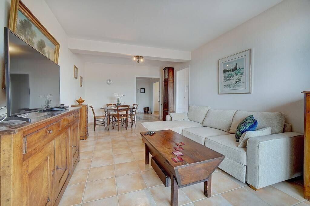 A living room with a sofa, coffee table, wooden cabinet, and a TV. There is a dining area with chairs and a table in the background.