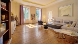 A spacious living room with a wooden floor, a white sofa, a black chair, and a coffee table. There are large windows with curtains, a painting on the wall, and a bookshelf with various items.