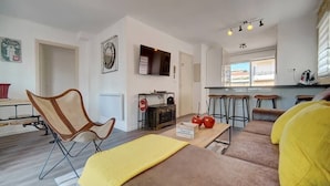 Een moderne woonkamer met een bank, een stoel, een salontafel en een televisie. Er is een keuken met krukken en een raam.