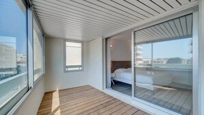 Een modern balkon met houten vloer, grote ramen en een schuifdeur naar een slaapkamer met een bed en nachtkastje.
